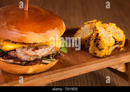 Hamburger de boeuf avec du fromage et de la laitue et de maïs grillé, servi sur plaque de bois sur la table en bois Banque D'Images