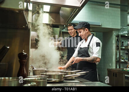 L'humeur positive. Vue latérale des deux joyeux et souriant en uniforme cuisiniers préparent une cuisine dans un restaurant de cuisine. Concept de préparation des aliments Banque D'Images