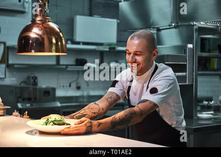 Au moment de servir. Young smiling chef de tatouages sur ses bras, en tablier noir tenant prêt plat dans une cuisine de restaurant moderne. Concept alimentaire Banque D'Images
