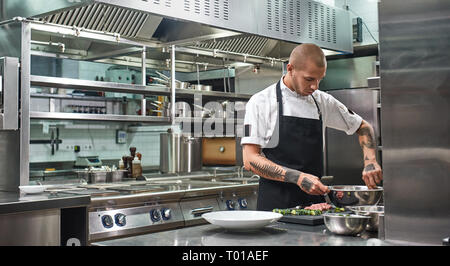 Presque prêt. Beau chef professionnel avec des tatouages sur ses bras un mélange salade dans un bol en métal tout en se tenant dans une cuisine. Cuisson Banque D'Images