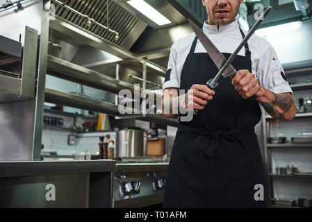 Chef professionnel. Jeune homme brutal en tablier avec plusieurs tatouages sur ses bras un affûtage couteau dans une cuisine de restaurant. Concept de cuisine Banque D'Images