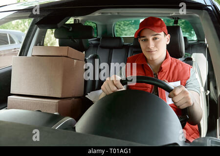 La distribution des colis postaux de l'homme en uniforme rouge conduit une voiture. Il est en train de regarder la caméra avec un sourire. Courrier postal Transport La livraison La livraison des colis aux clients. Service impressionnant. Banque D'Images