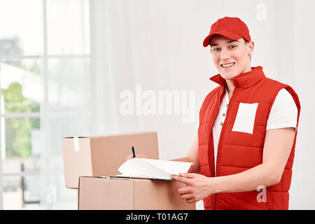 Portrait de l'homme en rouge livraison uniforme postal debout près de la pile de boîtes, l'étude de la liste des clients pour aujourd'hui. Friendly travailleur, le service de haute qualité. À l'intérieur. Banque D'Images