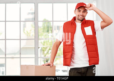 Cheerful postman porter du rouge uniforme postale est la livraison colis à un client. Il a fait son travail très bien. Il touche son chapeau et regardant la caméra avec un sourire. Friendly travailleur, le service de haute qualité. À l'intérieur. Intérieur lumineux. Banque D'Images