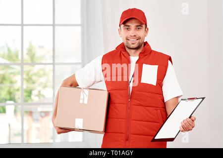 Cheerful postman porter du rouge uniforme postale est la livraison colis à un client. Il est titulaire d'un emballage en carton, dossier et regardant la caméra avec un sourire. Friendly travailleur, le service de haute qualité. À l'intérieur. Intérieur lumineux. Banque D'Images