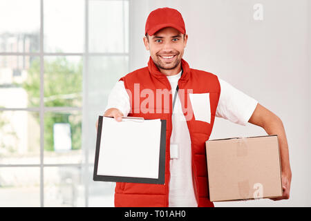 Cheerful postman porter du rouge uniforme postale est la livraison colis à un client. Il est titulaire d'un emballage en carton, en attente d'une signature à la livraison et regardant la caméra avec un sourire. Friendly travailleur, le service de haute qualité. À l'intérieur. Intérieur lumineux. Banque D'Images