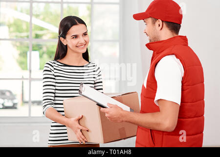 Cheerful postman porter du rouge uniforme postale est la livraison colis à un client satisfait. Il apporte en carton à son propriétaire. Dark-haired woman prend des colis, elle est heureuse de le recevoir. Friendly travailleur, le service de haute qualité. À l'intérieur. Intérieur lumineux. Banque D'Images