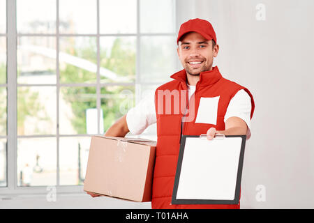 Cheerful postman porter du rouge uniforme postale est la livraison colis à un client. Il est titulaire d'un emballage en carton, en attente d'une signature à la livraison et regardant la caméra avec un sourire. Friendly travailleur, le service de haute qualité. À l'intérieur. Intérieur lumineux. Banque D'Images