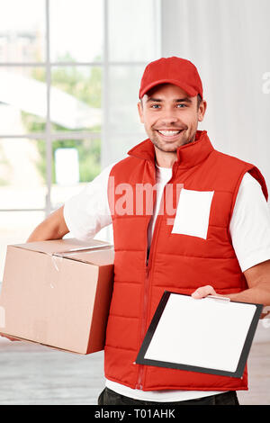 Cheerful postman porter du rouge uniforme postale est la livraison colis à un client. Il est titulaire d'un emballage en carton, en attente d'une signature à la livraison et regardant la caméra avec un sourire. Friendly travailleur, le service de haute qualité. À l'intérieur. Intérieur lumineux. Banque D'Images