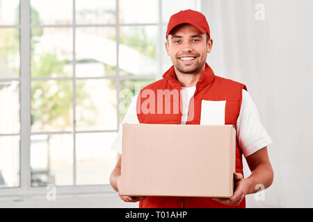 Cheerful postman porter du rouge uniforme postale est la livraison colis à un client. Il est titulaire en carton et regardant la caméra avec un sourire. Friendly travailleur, le service de haute qualité. À l'intérieur. Intérieur lumineux. Banque D'Images