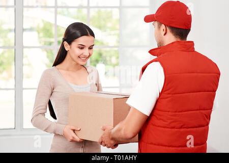 Cheerful postman porter du rouge uniforme postale est la livraison colis à un client satisfait. Il apporte en carton à son propriétaire. Dark-haired woman prend des colis, elle est heureuse de le recevoir. Friendly travailleur, le service de haute qualité. À l'intérieur. Intérieur lumineux. Banque D'Images