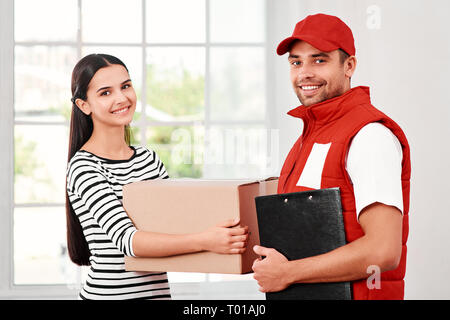 Cheerful postman porter du rouge uniforme postale est la livraison colis à un client satisfait. Il est titulaire d'receing rapport de livraison après un signe de la part du client. Dark-haired woman détient en carton et regardant la caméra avec un sourire. Friendly travailleur, le service de haute qualité. À l'intérieur. Intérieur lumineux. Banque D'Images