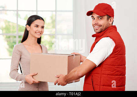 Cheerful postman porter du rouge uniforme postale est la livraison colis à un client satisfait. Il est en train de regarder l'appareil photo, alors qu'elle se penche sur lui. Ils détiennent en carton et souriant. Friendly travailleur, le service de haute qualité. À l'intérieur. Intérieur lumineux. Banque D'Images