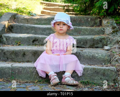 Mignon bébé fille aux yeux de graphite est assis pensivement sur les anciennes marches de pierre couverts de mousse Banque D'Images