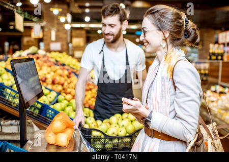 Beau d'aider les jeunes travailleurs en uniforme femme client pour peser sur la balance des pommes dans le supermarché Banque D'Images