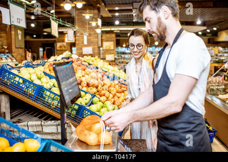 Beau d'aider les jeunes travailleurs en uniforme femme client pour peser sur la balance des pommes dans le supermarché Banque D'Images