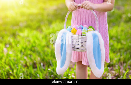 Les enfants avec des oeufs panier et oreilles de lapin sur la chasse aux œufs de Pâques dans le jardin de printemps ensoleillé. Petite fille à la recherche de bonbons colorés et des œufs en chocolat avec le rabbin Banque D'Images