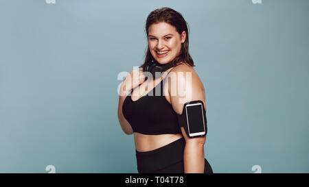 Portrait de jeune femme en surpoids dans les vêtements de sport. Femme posant dans sportswear sur fond gris avec téléphone mobile sur brassard et un casque à la Banque D'Images
