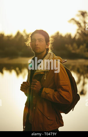 Homme portant une veste et un sac à dos debout dehors au bord d'un lac. voyageur debout à l'extérieur regardant loin avec le soleil en arrière-plan. Banque D'Images