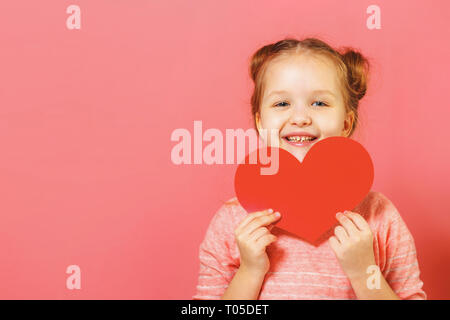 Closeup portrait of a cute little girl avec brioches de cheveux sur un fond rose. Child holding heart Banque D'Images