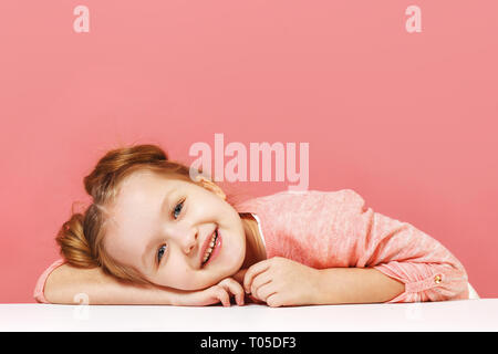 Closeup portrait of a cute little girl avec cheveux buns sur fond rose. Smiling enfant d'âge préscolaire mis sa tête sur la table Banque D'Images