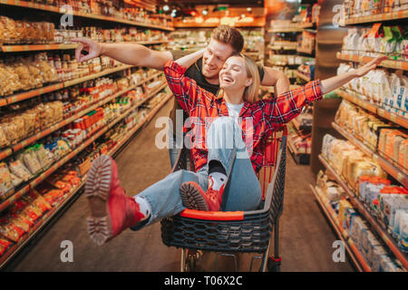 Jeune couple en épicerie. Femme s'asseoir dans le chariot et s'amuser. Homme debout derrière et le point sur la tablette avec produits. Couple heureux d'avoir du plaisir. Banque D'Images