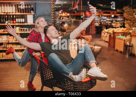 Jeune couple en épicerie. L'homme assis dans le chariot ludique et vague avec les mains. Happy blonde woman se tenir derrière. Elle pousser trolley plus loin. Banque D'Images
