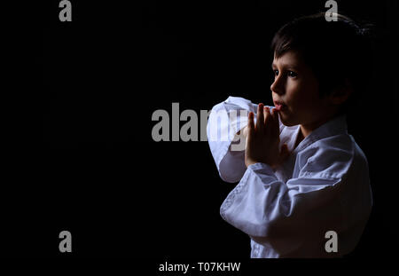 Peu de karaté kid en kimono blanc isolé sur fond sombre. Portrait of boy prêt pour les arts martiaux lutte. La lutte contre l'enfant à la formation de judo. Meilleures con Banque D'Images