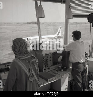 Années 60, le contrôle du trafic aérien, l'aéroport de Riyad, Arabie saoudite, deux contrôleurs au sol, l'une en robe arabe traditionnelle, debout dans leur kiosque d'observation à la recherche sur la voie de circulation et la piste pour visual vue de l'avion à réaction. Banque D'Images