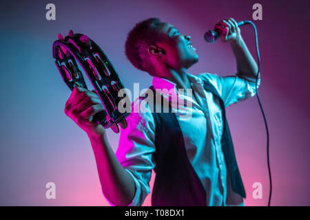 Le musicien de jazz afro-américain beau jouant du tambourin et en chantant dans le microphone en studio sur un fond de néon. Music concept. Les jeunes gars attrayant joyeuse improvisation. Close-up portrait rétro. Banque D'Images