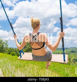 Happy female traveller swinging sur balançoire en bois, appréciant les vacances d'été entre les terrasses de riz vert immaculé. Banque D'Images