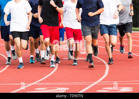 Les garçons du secondaire en cours d'exécution dans un grand groupe sur une piste de cross-country rouge au cours de la pratique. Banque D'Images