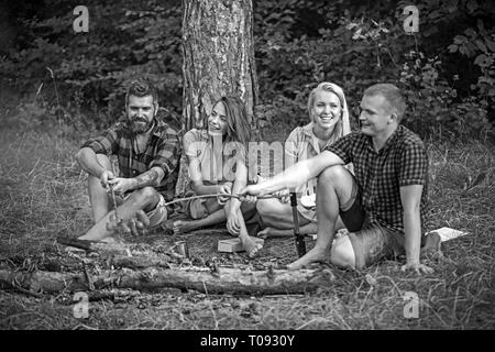 Deux couples camping dans les bois. Saucisses à frire les gars sur le feu. Smiling friends sitting autour de feu de camp le soir Banque D'Images