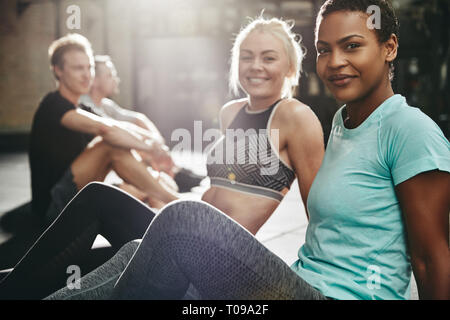 Deux jeunes femmes en souriant sportswear assis ensemble sur le plancher d'une salle de sport après une séance avec deux amis masculins Banque D'Images