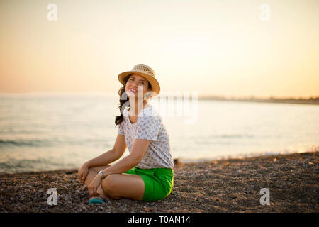 Portrait of a young woman sitting cross legged sur la plage au coucher du soleil. Banque D'Images