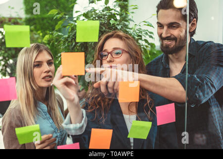 Les créatifs sont debout et discuter au bureau derrière un mur de verre avec des notes adhésives et à un mur de post-it. Banque D'Images