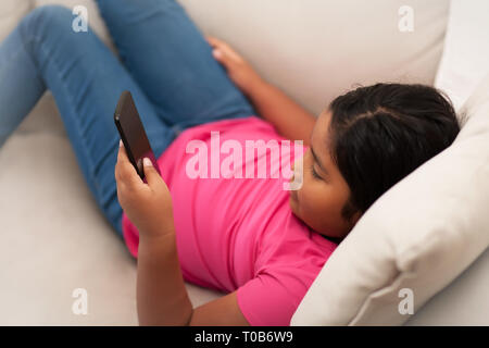 Jeune fille assise avec les jambes allongées sur un canapé à l'aide d'un téléphone à texte ou afficher le contenu en ligne sans supervision. Banque D'Images