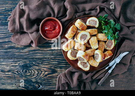 Rouleaux de saucisses sur une plaque de faïence sur une table rustique en bois foncé avec chiffon brun et de la sauce tomate dans un bol, finger food, cuisine anglaise traditionnelle, voir fr Banque D'Images