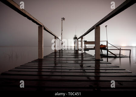 Silhouette d'un homme debout sur un quai en bois dans le noir, fixant le vide d'un lac avec feux lumineux sur le côté opposé Banque D'Images