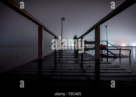 Silhouette d'un homme debout sur un quai en bois dans le noir, fixant le vide d'un lac avec feux lumineux sur le côté opposé Banque D'Images