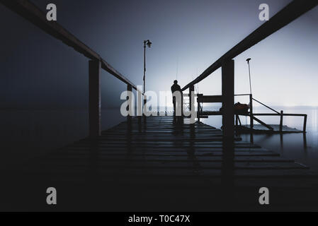 Silhouette d'un homme debout sur un quai en bois dans le noir, fixant le vide d'un lac avec feux lumineux sur le côté opposé Banque D'Images