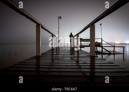 Silhouette d'un homme debout sur un quai en bois dans le noir, fixant le vide d'un lac avec feux lumineux sur le côté opposé Banque D'Images