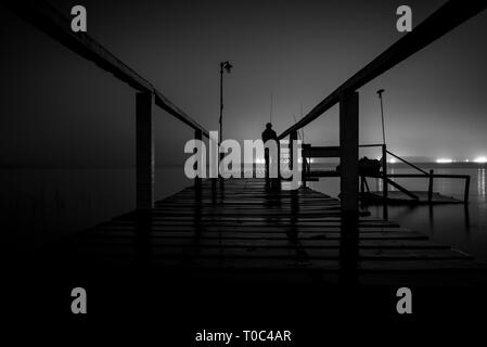 Silhouette d'un homme debout sur un quai en bois dans le noir, fixant le vide d'un lac avec feux lumineux sur le côté opposé Banque D'Images