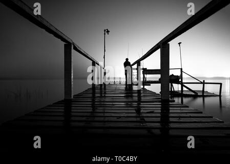 Silhouette d'un homme debout sur un quai en bois dans le noir, fixant le vide d'un lac avec feux lumineux sur le côté opposé Banque D'Images