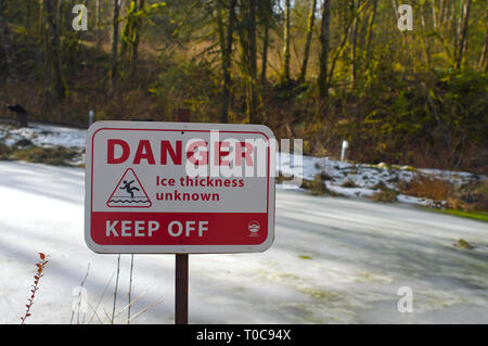 L'épaisseur de la glace danger inconnu Garder hors panneau d'avertissement à côté d'un étang couvert de glace avec des arbres en arrière-plan. Banque D'Images