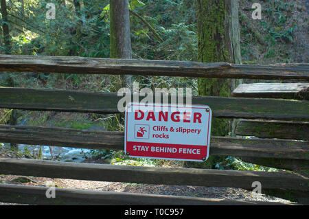 Danger Falaises & Roches glissantes restent derrière clôture Panneau d'avertissement sur un rail vertical clôture avec arbres de derrière. Banque D'Images