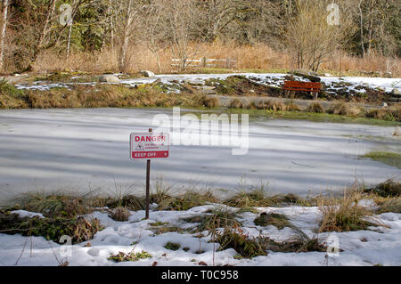 Panneau d'avertissement "danger lecture inconnue de l'épaisseur de la glace Garder Off' à côté d'un étang couvert de glace avec un banc de l'autre côté. Banque D'Images