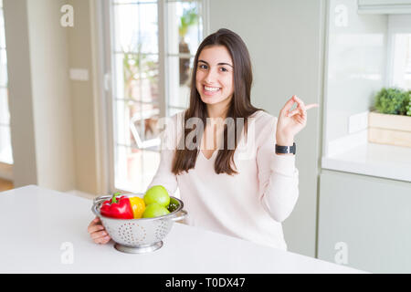 Belle jeune femme à l'aide d'une passoire pour laver et nettoyer légumes très heureux pointant avec la main et le doigt sur le côté Banque D'Images