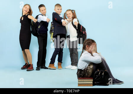 Petite fille assise seule sur le plancher et les souffrances d'un acte d'intimidation alors que les enfants se moquer. Triste jeune écolière assis sur studio contre fond bleu. Banque D'Images