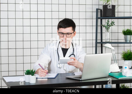 Médecin homme en blouse blanche avec stéthoscope sur son cou sitting at table pensant sur prescription, d'écrire quelque chose vers le bas, avec la boîte de médecine Banque D'Images